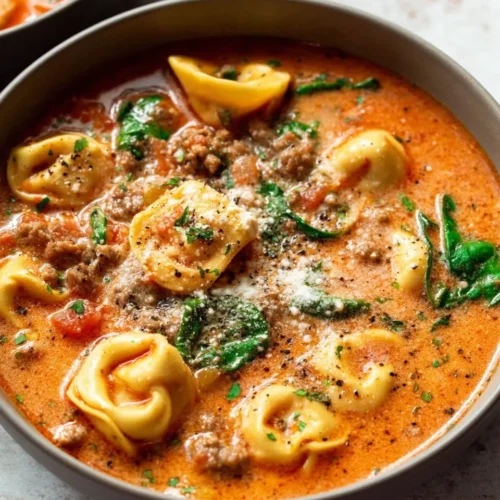 A close-up overhead view of a beige bowl filled with creamy Slow Cooker Tortellini Soup with Sausage, featuring plump cheese tortellini, browned sausage crumbles, wilted spinach, and a sprinkling of grated Parmesan cheese.