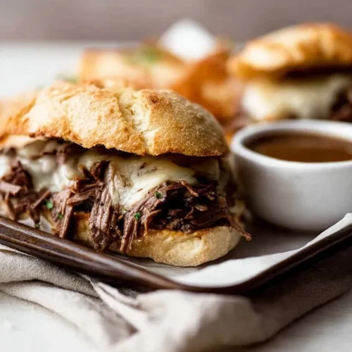 Close-up of a Slow Cooker French Dip sandwich on a toasted crusty roll, filled with tender shredded beef and melted white cheese, served alongside a white ramekin of rich au jus dipping sauce on a lined tray.