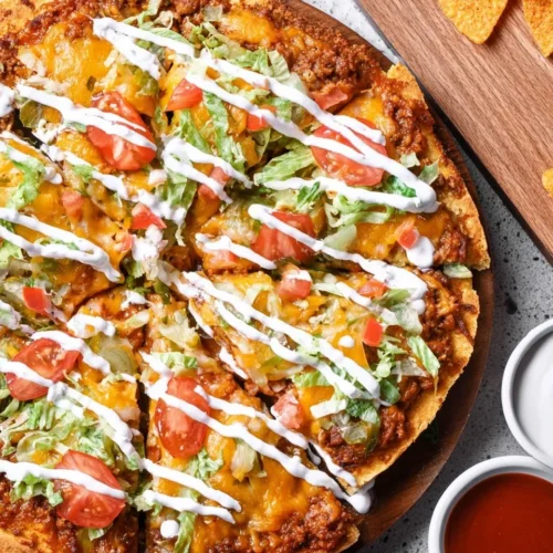 Top-down view of a sliced taco pizza featuring seasoned taco meat pizza topping, melted cheddar cheese, shredded lettuce, and diced tomatoes, drizzled with sour cream on a wooden board.