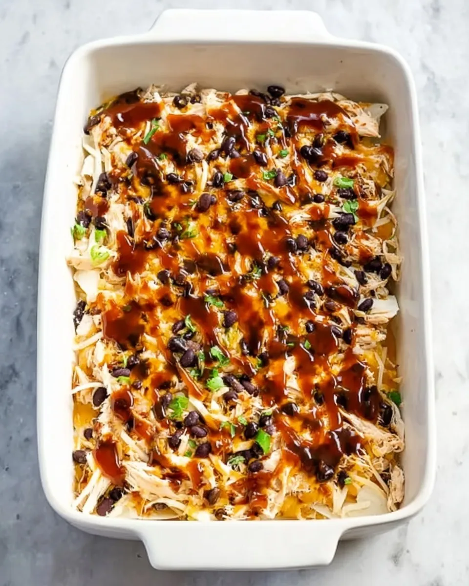 Top-down view of chicken enchilada casserole assembly showing shredded chicken and black bean filling drizzled with red enchilada sauce in a white baking dish.