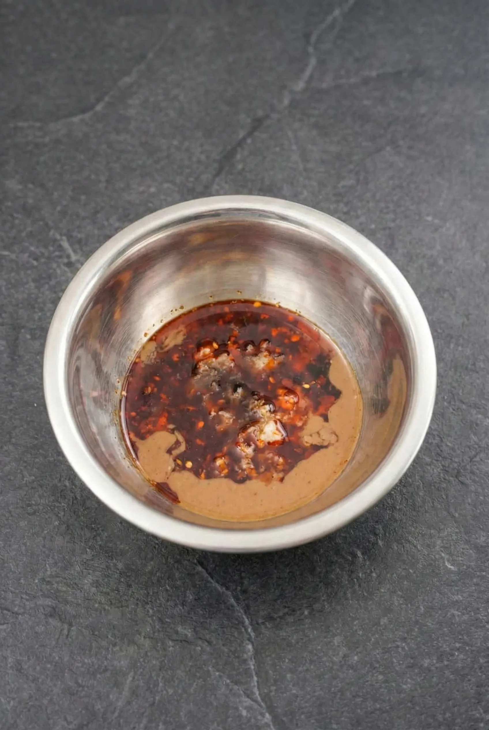 Overhead view of a stainless steel bowl containing unmixed sesame sauce ingredients, including creamy sesame paste, vibrant red chili oil, soy sauce, and minced garlic against a dark stone background.