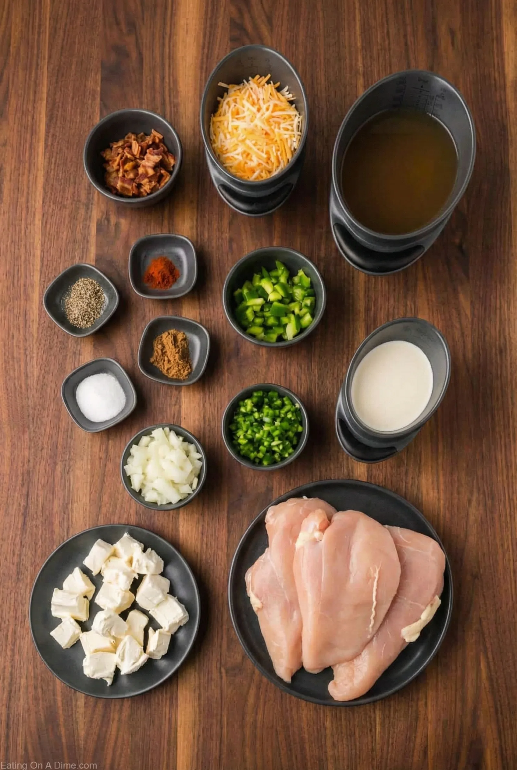 Crockpot Jalapeño Popper Chicken Soup ingredients Top-down view of the ingredients for Crockpot Jalapeño Popper Chicken Soup arranged on a dark wooden table, including raw chicken breasts, cubed cream cheese, shredded cheese, chicken broth, heavy cream, chopped jalapeños, green peppers, onions, bacon bits, and spices.
