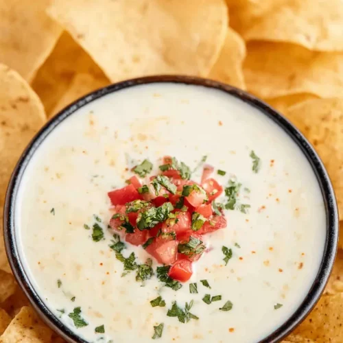 Top-down close-up view of a bowl of creamy Mexican white cheese dip topped with fresh pico de gallo and surrounded by crispy tortilla chips.