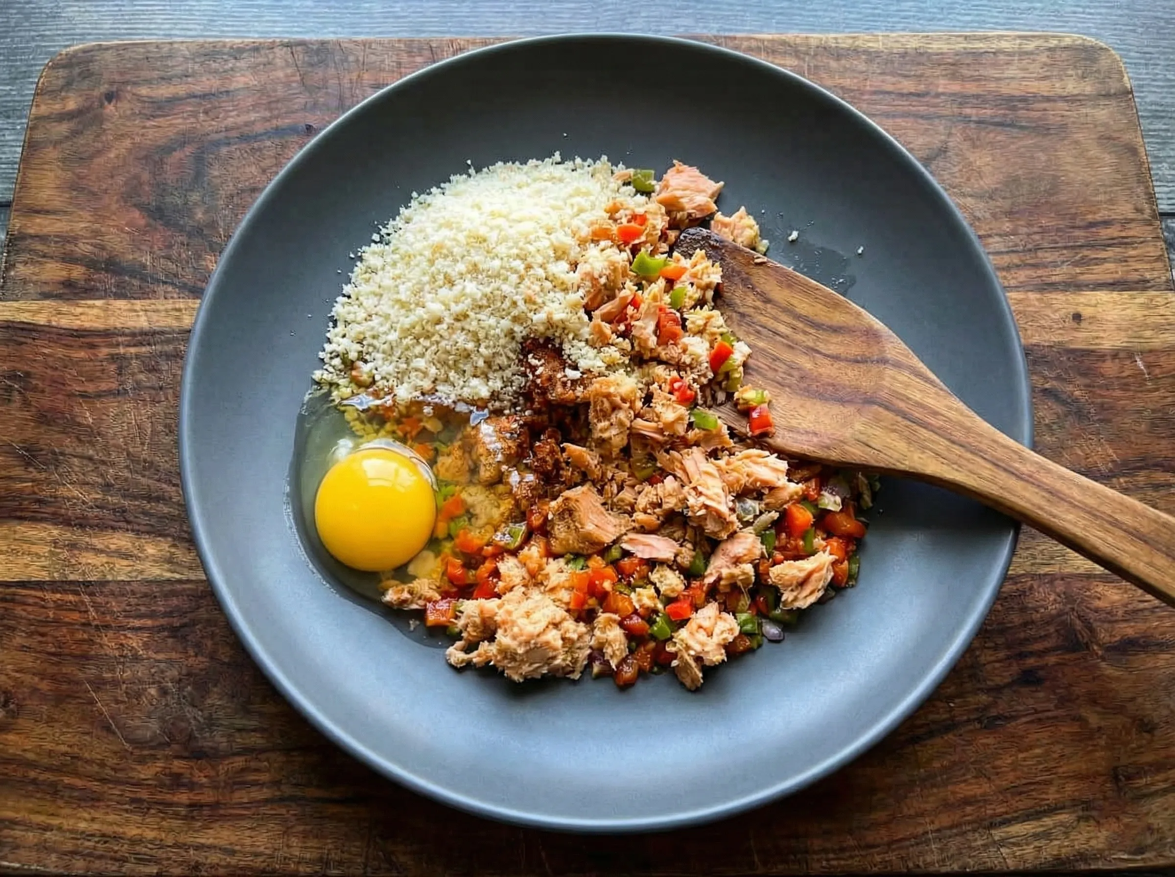 A dark mixing bowl containing ingredients for spicy salmon cakes, including canned salmon, panko breadcrumbs, a raw egg, and diced peppers with a wooden spoon.