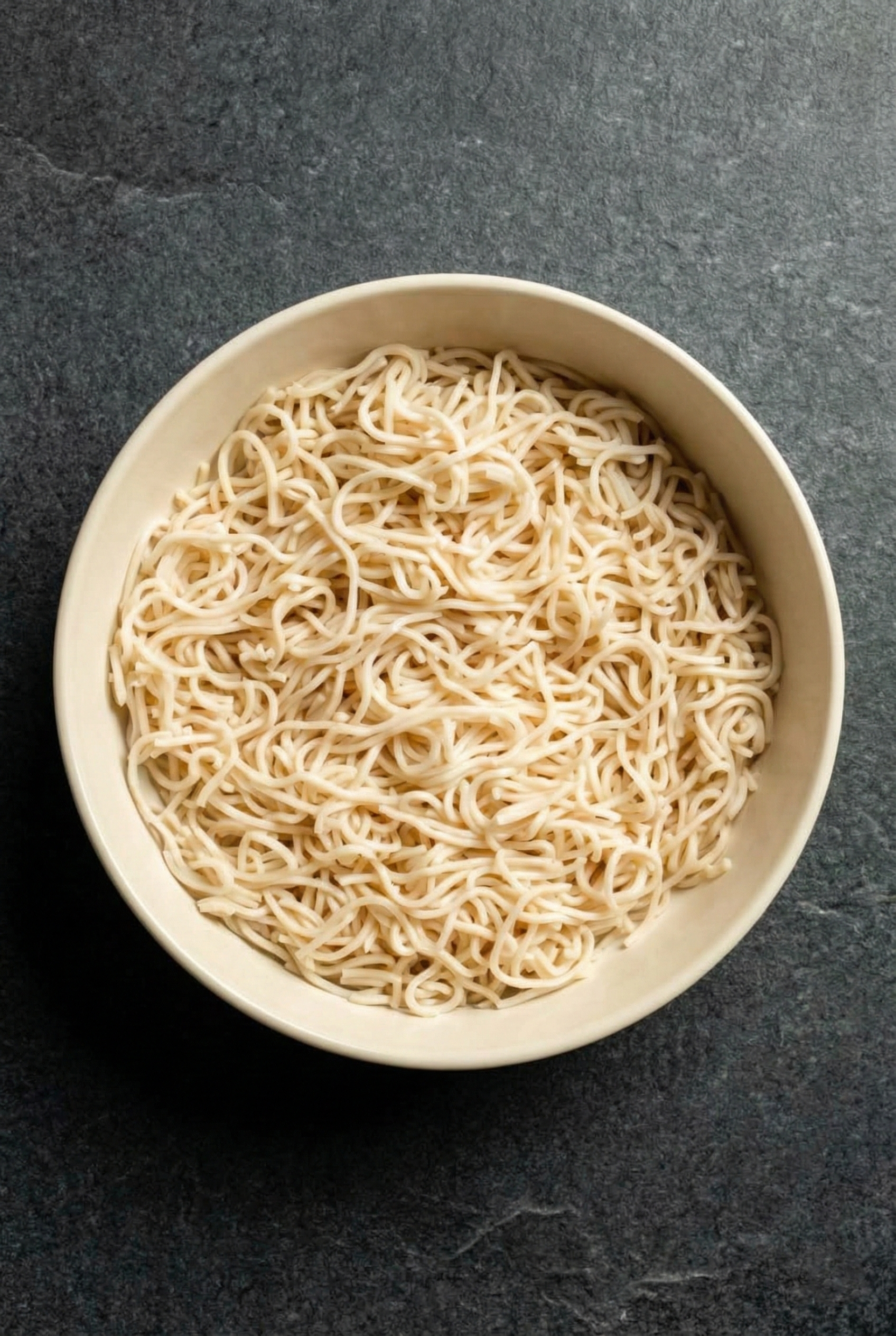 Top-down view of plain cooked thin wheat noodles in a beige bowl, rinsed and cooled, ready to be transformed into cold sesame noodles.
