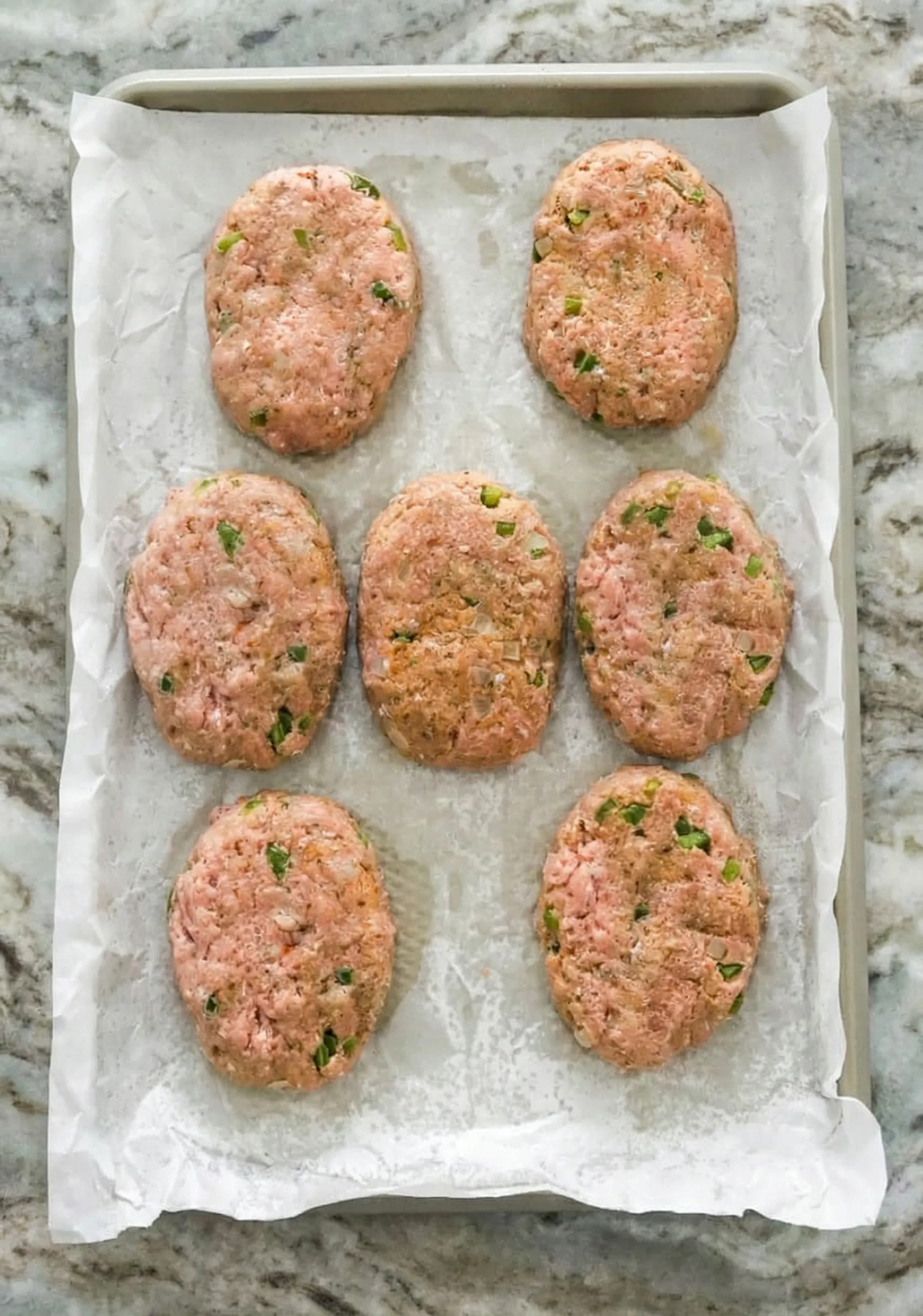 mini turkey meatloaves shaped into ovals on a parchment Overhead view of 8 raw mini turkey meatloaves shaped into ovals on a parchment-lined baking sheet, showing visible flecks of green herbs and onions.