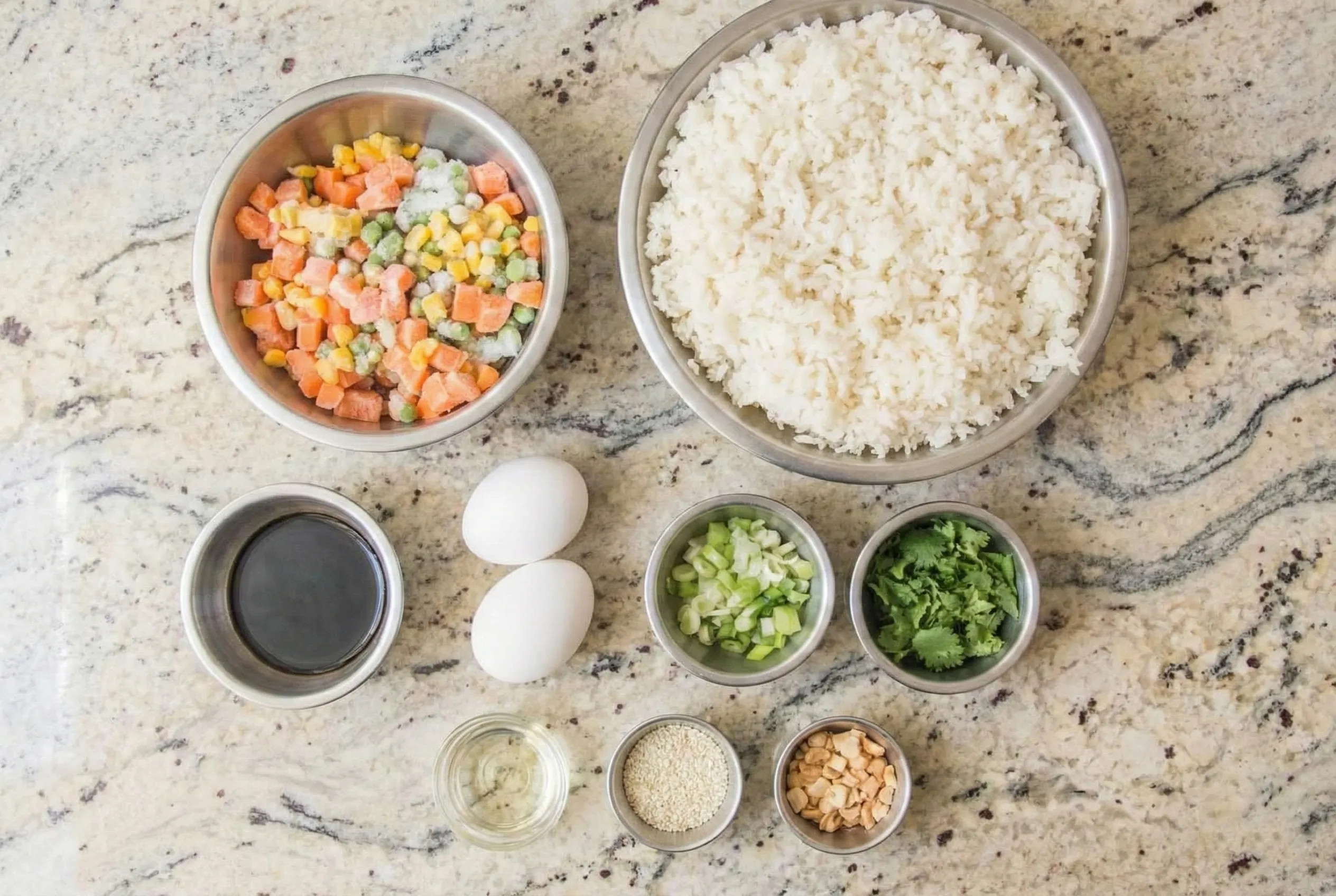 day old rice fried rice ingredients Flat lay of day old rice fried rice ingredients including a large bowl of cooked rice, frozen mixed vegetables, fresh eggs, soy sauce, and scallions arranged on a marble counter.