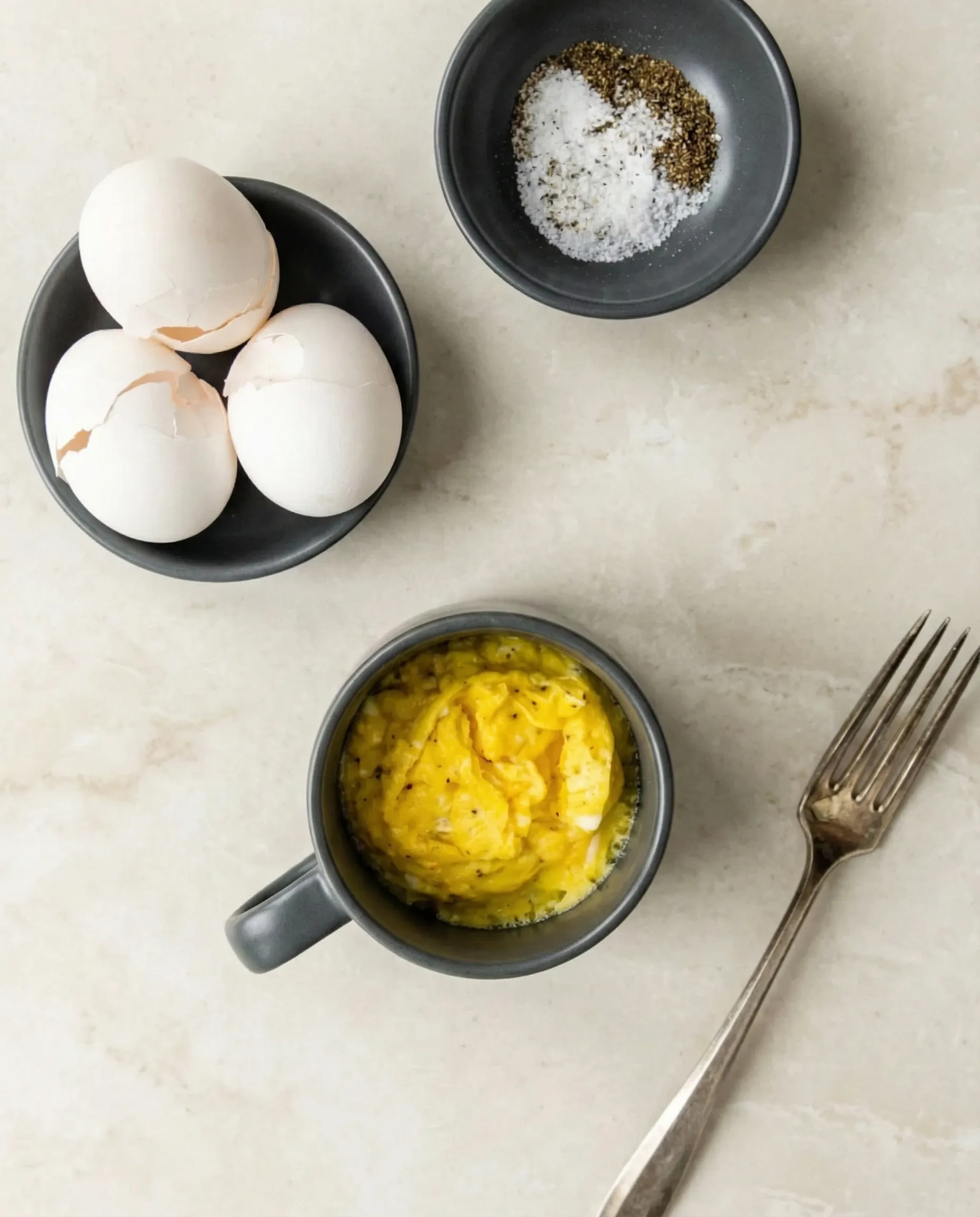 ingredients for microwave scrambled eggs Overhead flat lay showing ingredients for microwave scrambled eggs, including cracked eggshells, a bowl of salt and pepper, and fluffy cooked eggs in a grey mug with a silver fork.