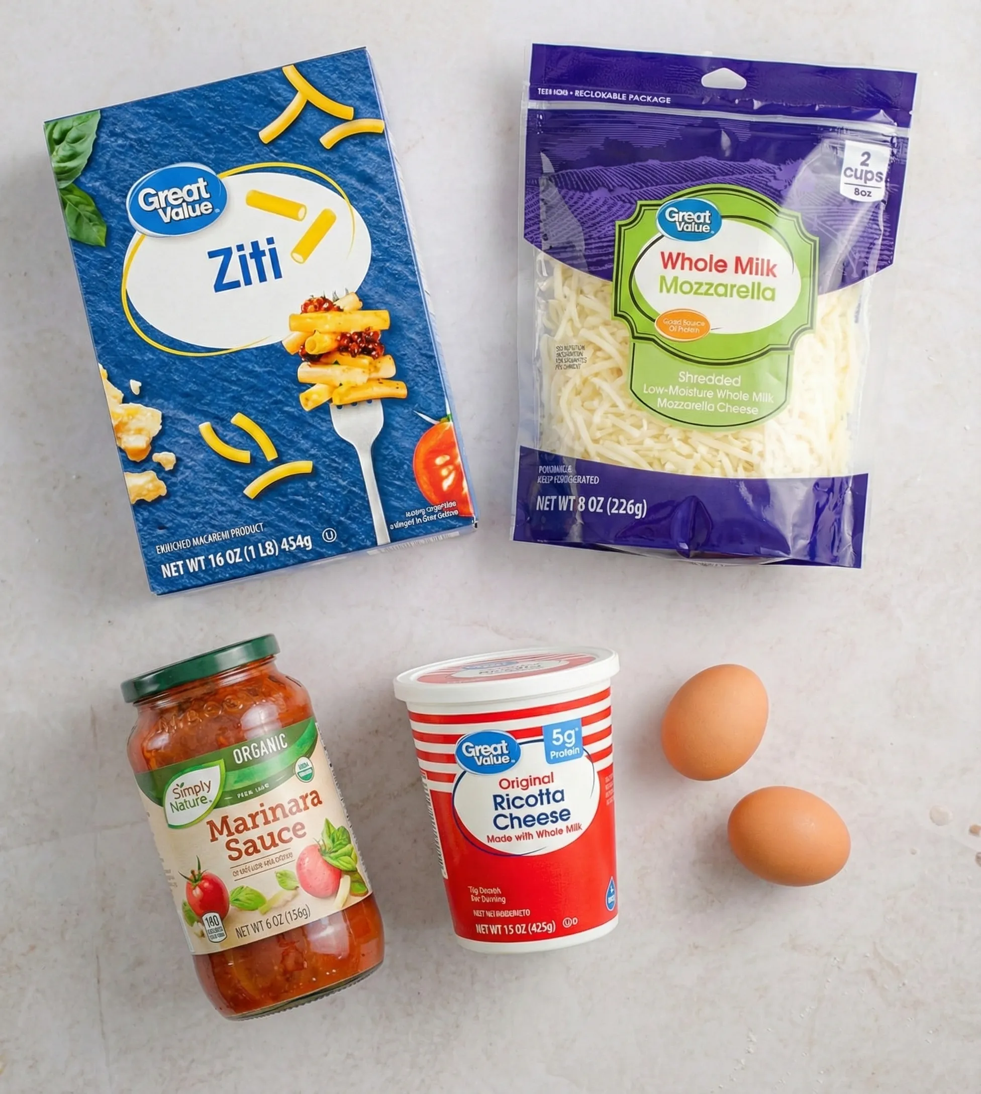 Overhead flat lay of ingredients for 5-ingredient cheesy baked ziti: a box of Great Value ziti pasta, a bag of shredded mozzarella cheese, a jar of Simply Nature marinara sauce, a container of ricotta cheese, and two brown eggs on a light marble surface.
