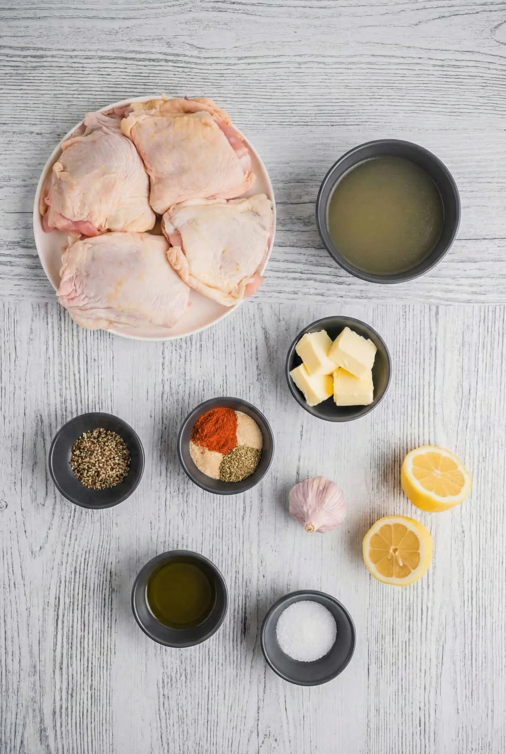 Fresh ingredients gathered for a Chicken Thigh Overhead flat lay of recipe ingredients on a white wooden table, featuring raw chicken thighs on a plate, a bowl of broth, cubed butter, fresh lemon halves, a whole garlic bulb, olive oil, and small bowls of spices including paprika, herbs, salt, and pepper.