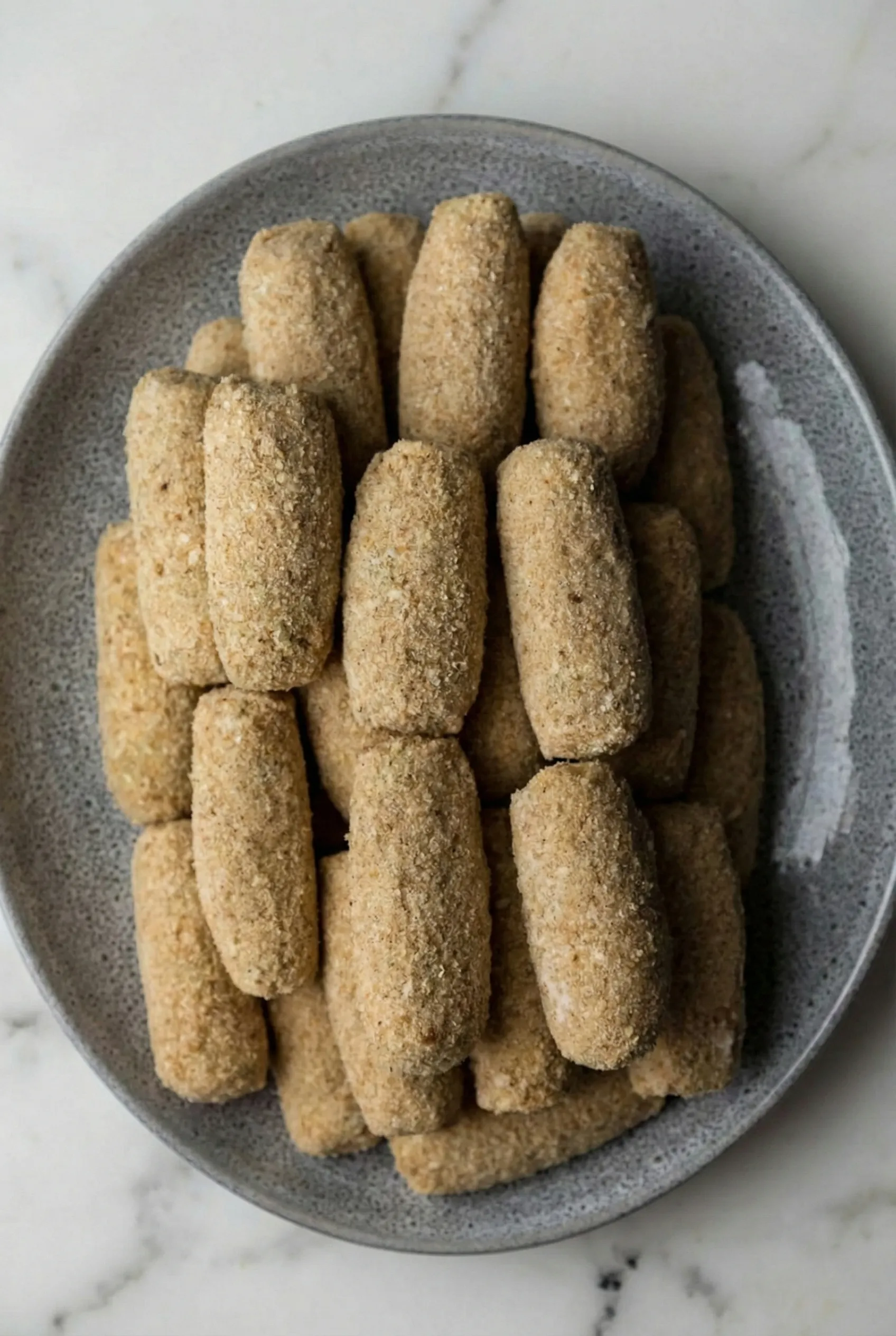 grey plate stacked high with uncooked mashed potato croquettes Top-down view of a grey plate stacked high with uncooked mashed potato croquettes that have been shaped into cylinders and evenly coated in breadcrumbs, ready for frying.