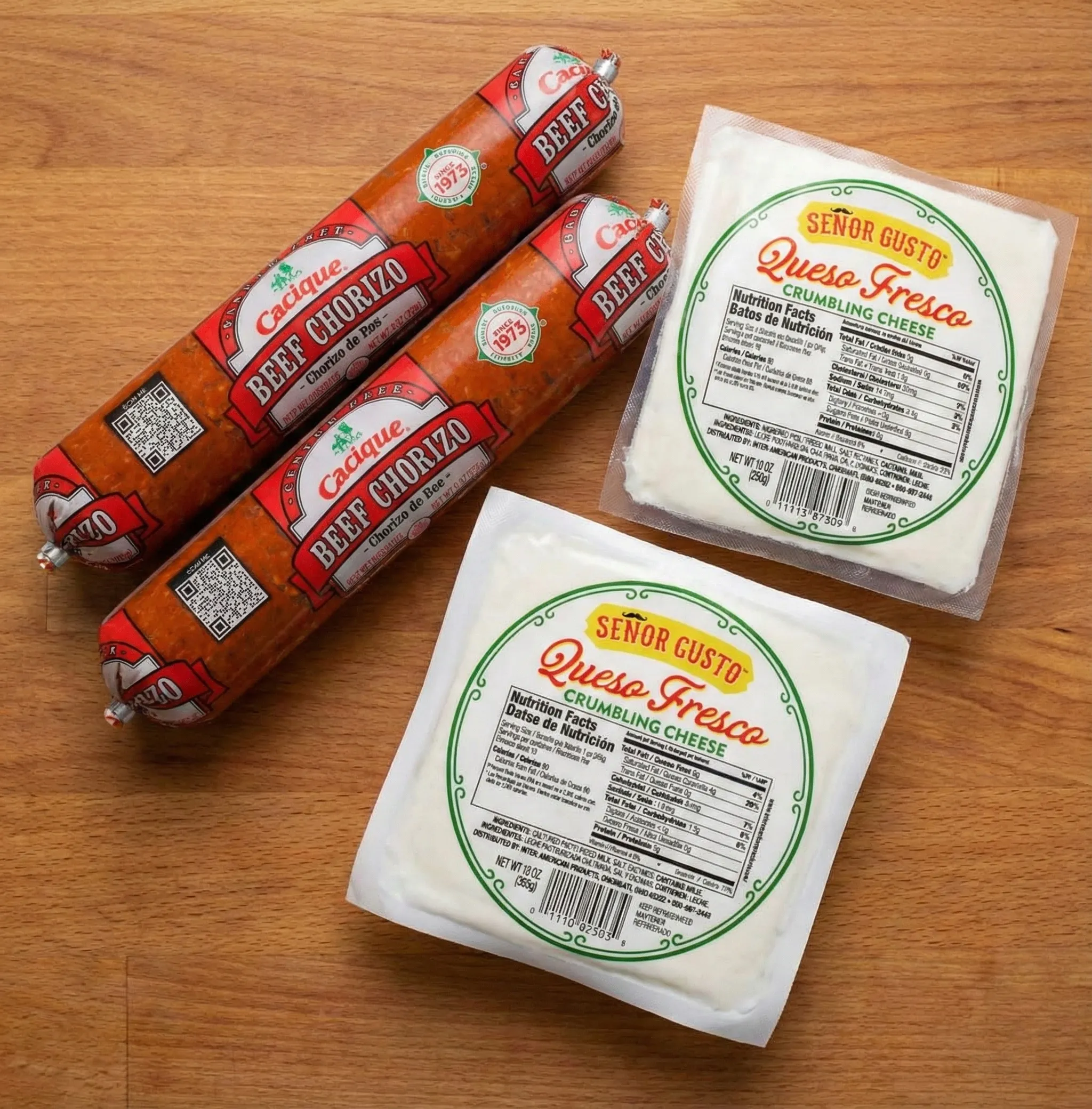 Ingredients for queso fundido recipe showing two tubes of Cacique beef chorizo and two blocks of Señor Gusto queso fresco cheese on a wooden table.