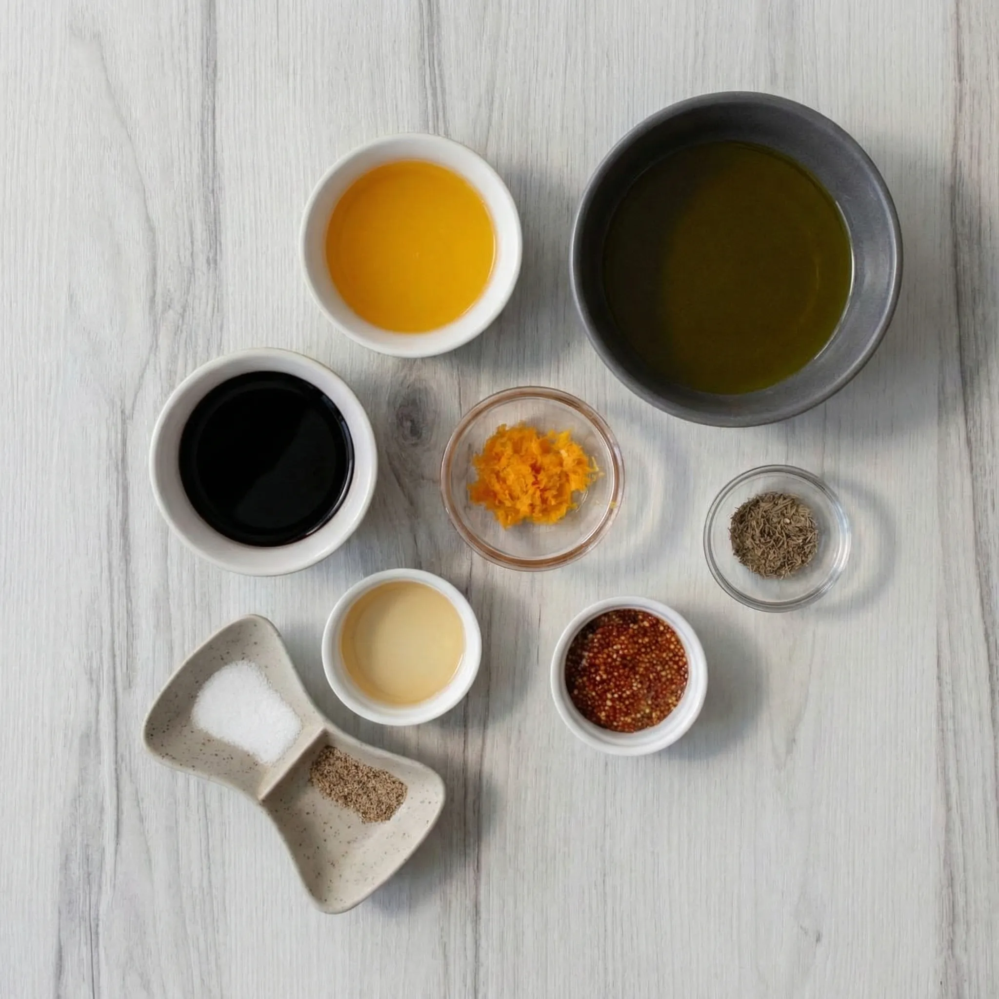 Overhead flat-lay of ingredients for Honey-Orange Balsamic Dressing, including olive oil, balsamic vinegar, orange juice, orange zest, whole grain mustard, honey, dried thyme, salt, and pepper arranged on a light wood surface.