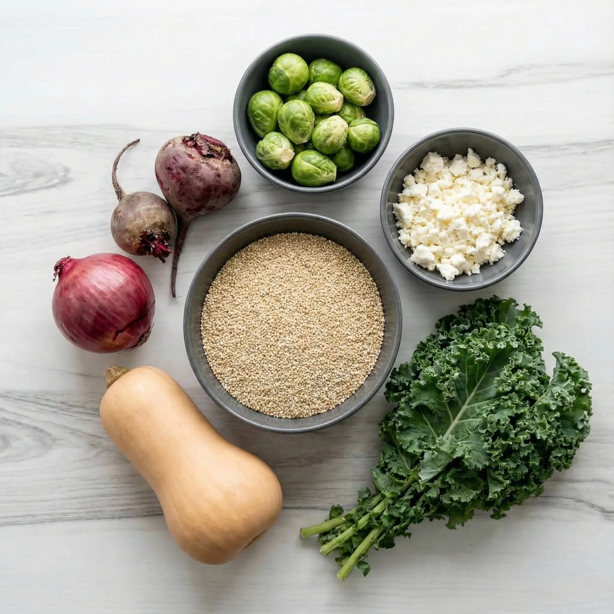 Overhead flat-lay view of fresh ingredients for Quinoa Salad with Roasted Vegetables, including a whole butternut squash, red onion, beets, Brussels sprouts, dried quinoa, crumbled feta, and fresh kale arranged on a white surface.