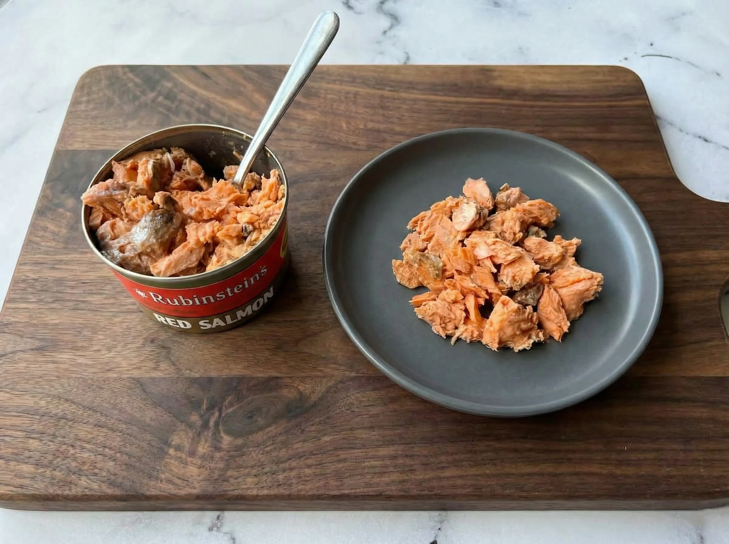 An open can of Rubinstein's Red Salmon with a spoon inside, sitting on a wooden board next to a small white plate of flaky canned salmon chunks.