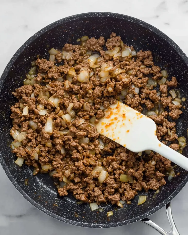 Overhead view of ground Italian sausage and chopped onions browning in a black speckled skillet with a white spatula.