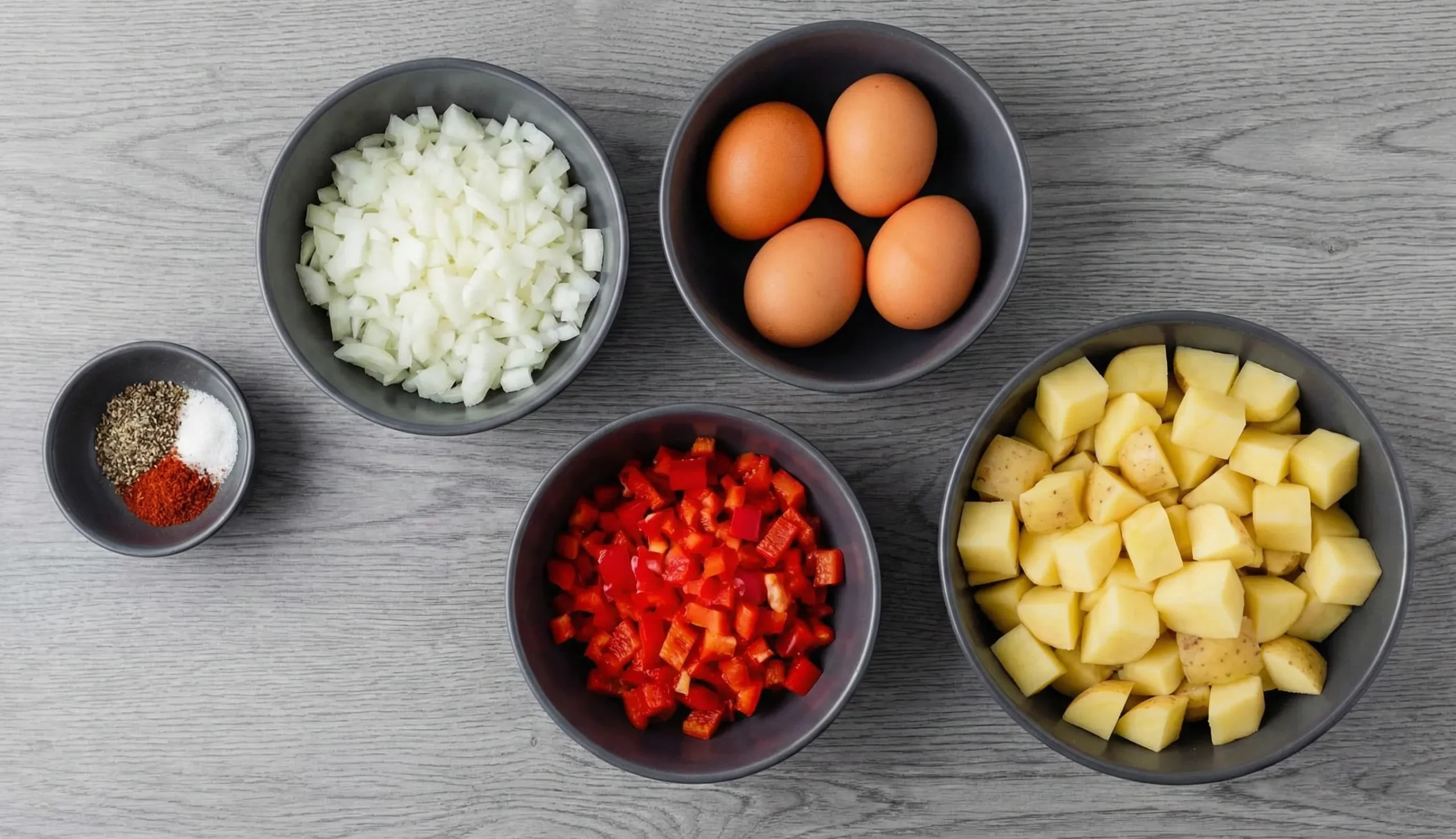 Flat lay of Potato and Egg Hash ingredients arranged in bowls: diced yellow potatoes, chopped white onions, diced red peppers, four brown eggs, and spices on a grey wood background.