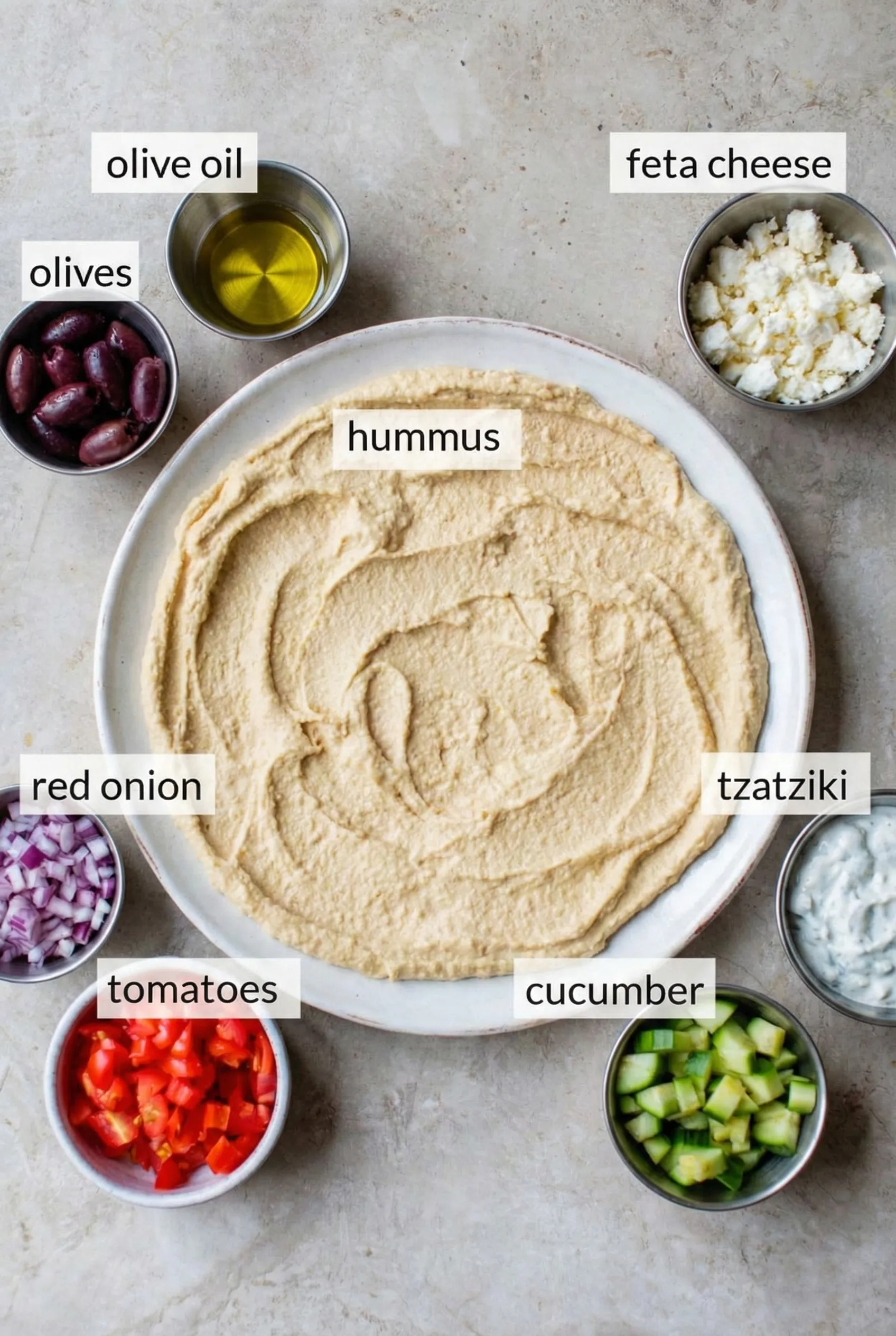 Mediterranean dip ingredients Top-down view of Mediterranean dip ingredients arranged on a marble counter, featuring a large plate with spread hummus surrounded by labeled bowls of diced red onion, tomatoes, cucumber, tzatziki, feta cheese, kalamata olives, and olive oil.