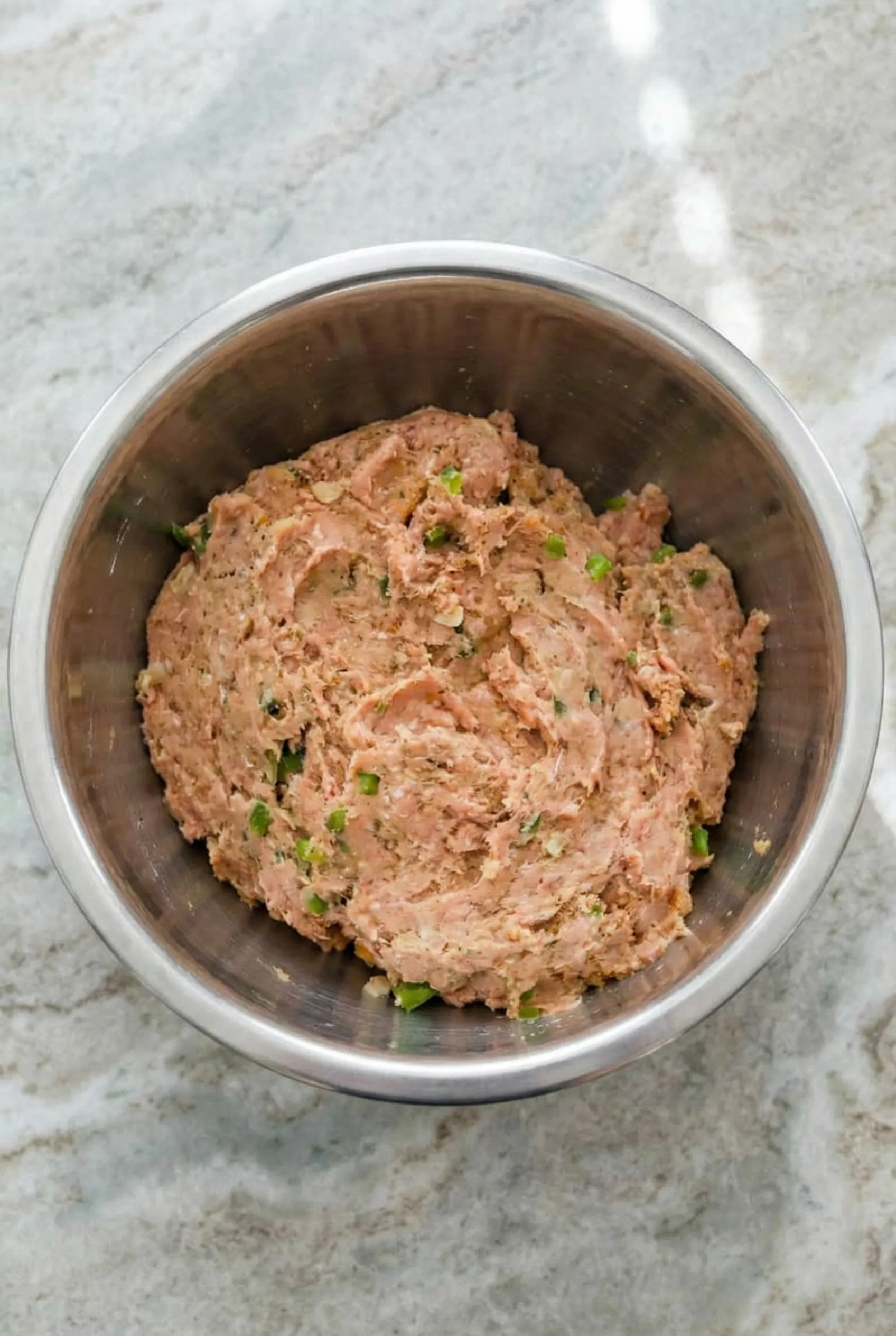 ground turkey meatloaf mixture A silver mixing bowl filled with raw ground turkey meatloaf mixture sitting on a marble counter, showing visible flecks of green onions and parsley evenly distributed throughout the meat.