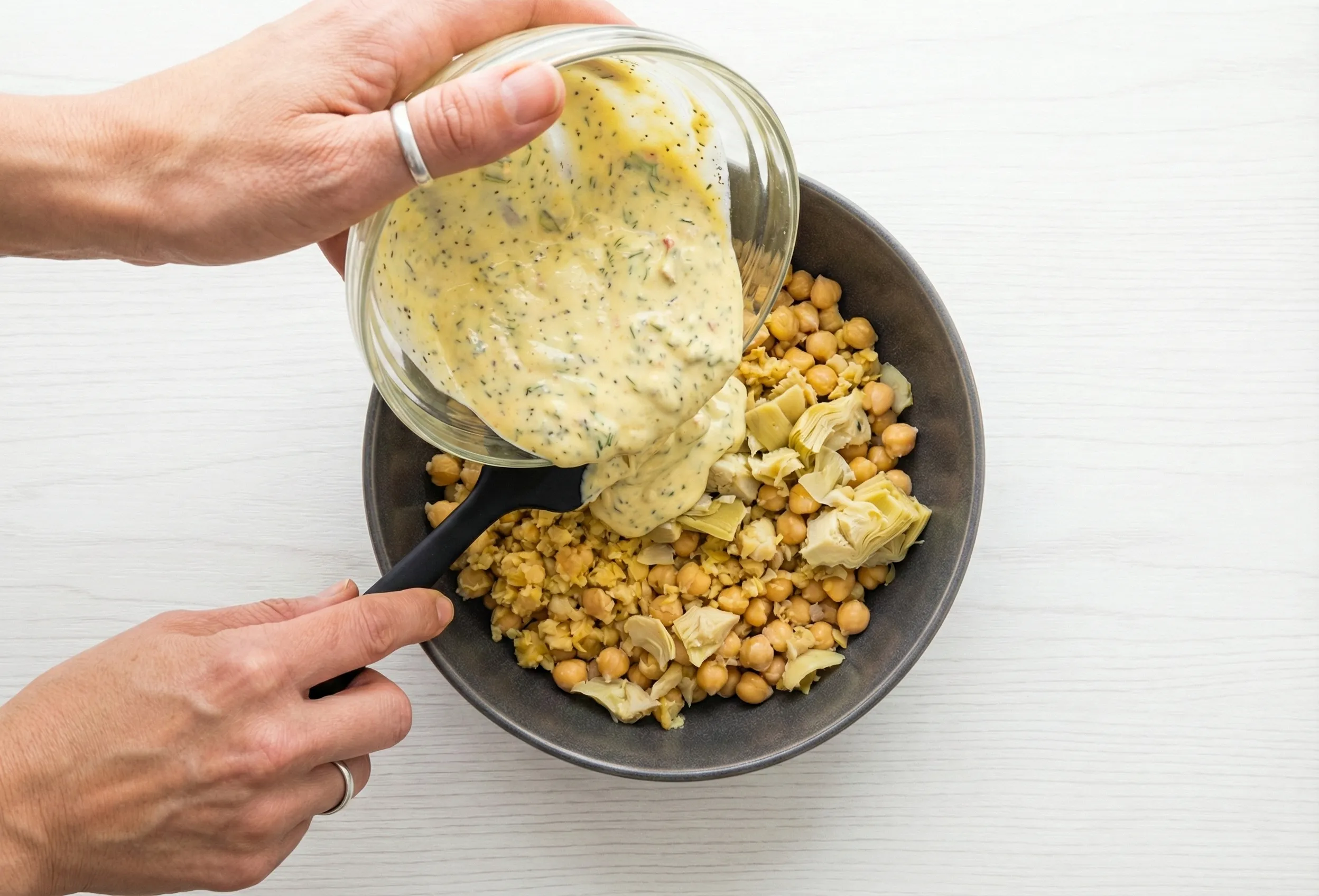 Hands pouring a creamy, herb-speckled yellow dressing from a glass bowl onto a mixture of mashed chickpeas in a dark grey bowl using a black spatula.