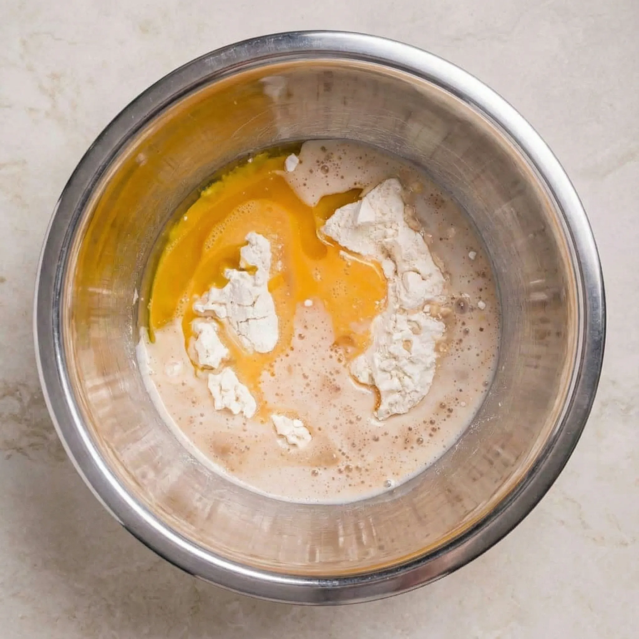 Stainless steel mixing bowl containing unmixed ingredients for small batch yeast rolls, including flour, beaten egg, and activated yeast liquid.