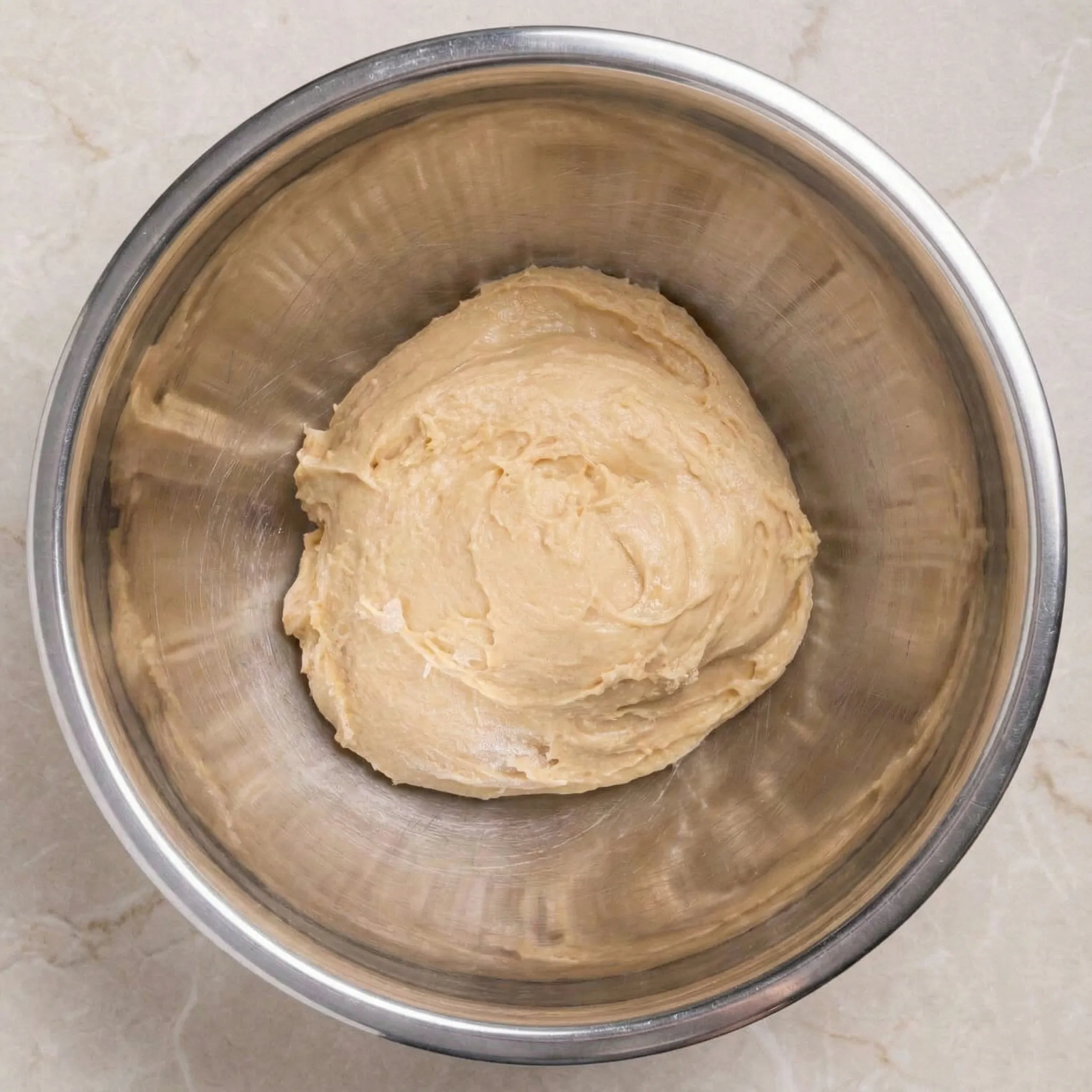 Overhead view of a stainless steel mixing bowl containing a ball of fully kneaded yeast dough that looks smooth and slightly tacky.