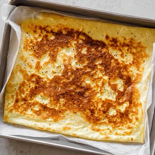 A large, golden-brown baked cottage cheese wrap sheet resting on parchment paper inside a baking pan. The surface has toasted brown spots and looks soft and pliable, fresh from the oven.
