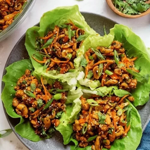 Four vibrant Asian chicken lettuce wraps arranged on a dark plate, garnished with shredded carrots, fresh cilantro, sliced green onions, and toasted sesame seeds.