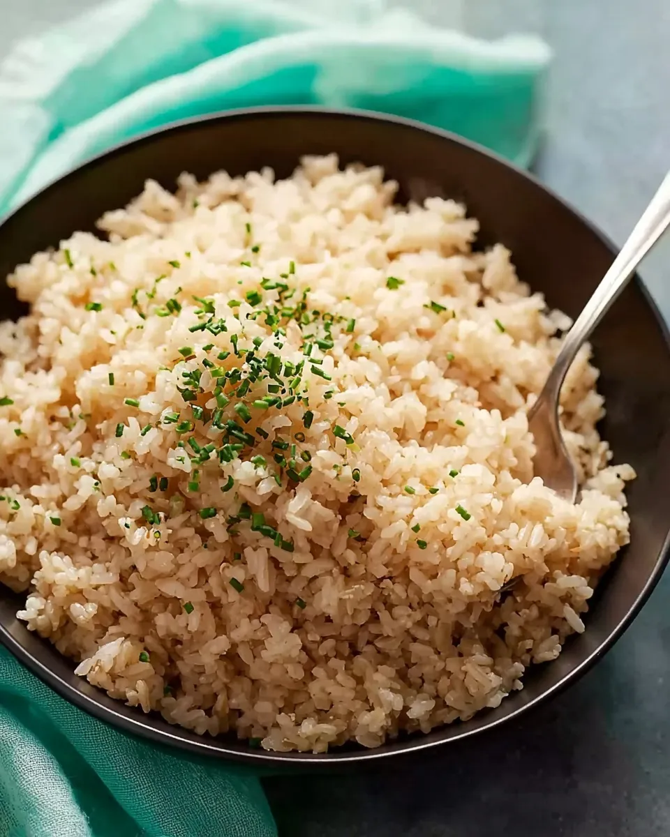 An overhead shot of a dark bowl piled high with perfectly cooked, fluffy brown rice, generously garnished with chopped fresh chives. A silver spoon is resting in the bowl, scooping up a serving.
