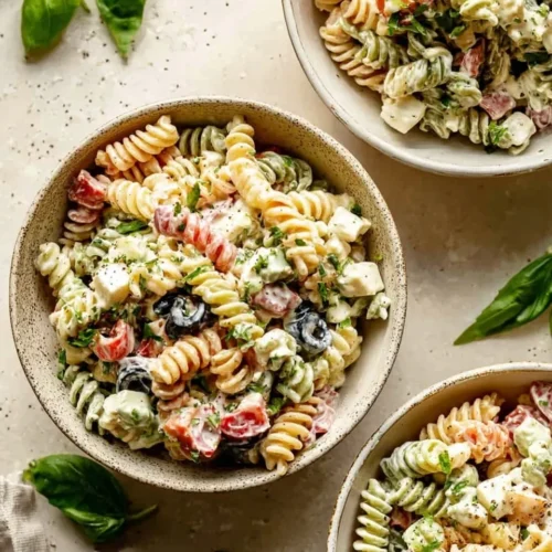 Three rustic ceramic bowls filled with individual servings of Creamy Cold Italian Pasta Salad, garnished with fresh herbs and showing tri-color rotini, diced mozzarella, and black olives.