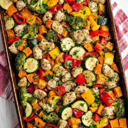 Overhead view of a large rimmed baking sheet filled with golden roasted chicken breast pieces, broccoli florets, diced sweet potatoes, red peppers, and yellow squash, set on a red and white striped kitchen towel.