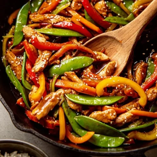 Close-up of a glossy chicken stir fry in a black cast-iron skillet featuring sliced red and yellow bell peppers, snap peas, and sesame seeds, with a wooden spoon and bowls of rice and red pepper flakes in the background.