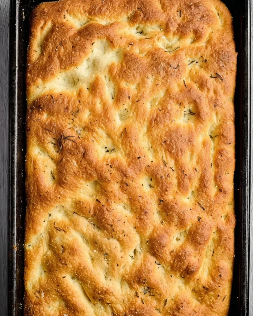 Top-down view of a freshly baked sheet pan focaccia in a dark rectangular baking sheet, featuring a golden-brown dimpled crust topped with dried rosemary and flaky sea salt.