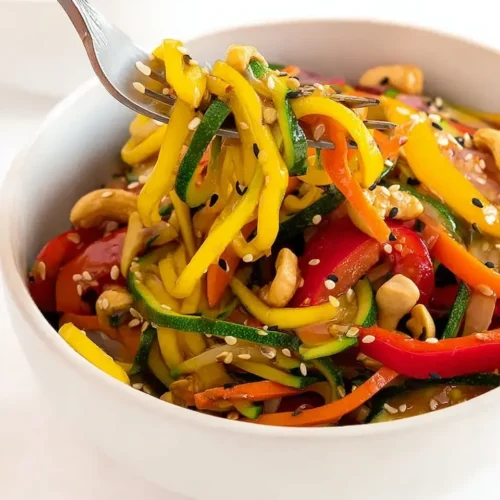 Close-up of a fork lifting a generous bite of Zucchini Noodle Stir-Fry from a white bowl, showing glossy spiralized zucchini, red bell peppers, roasted cashews, and a sprinkle of black and white sesame seeds.