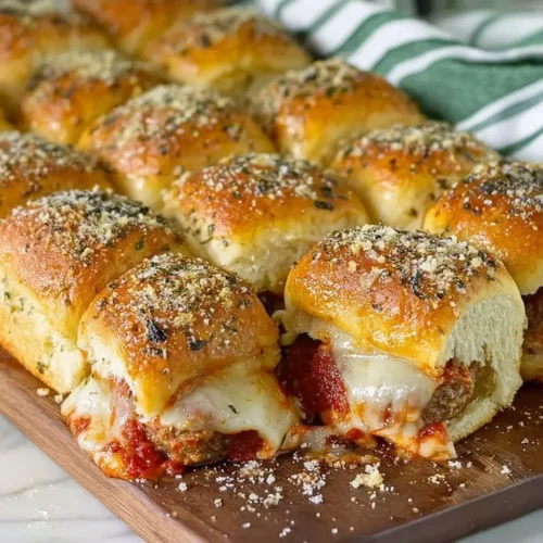 A close-up view of a dozen baked meatball sliders arranged on a wooden serving board. The golden-brown Hawaiian roll tops are glistening with garlic butter and sprinkled with herbs and Parmesan. Rich marinara sauce and melted white cheese are oozing out from the sides of the sliders.