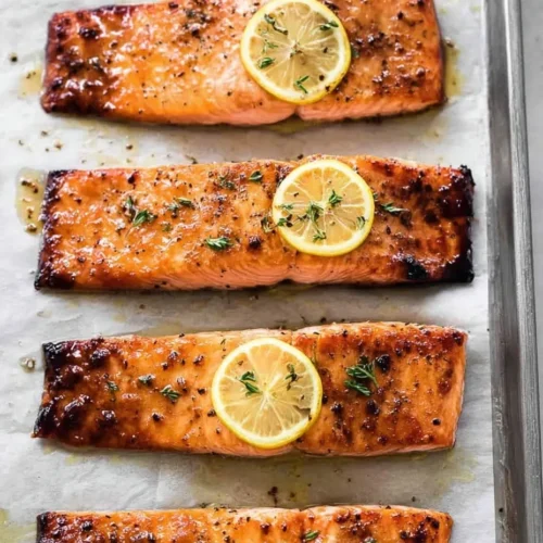 Four baked salmon filets on a parchment-lined baking sheet, showing a glossy maple mustard glaze, caramelized edges, fresh thyme sprigs, and a bright lemon slice on top of each.