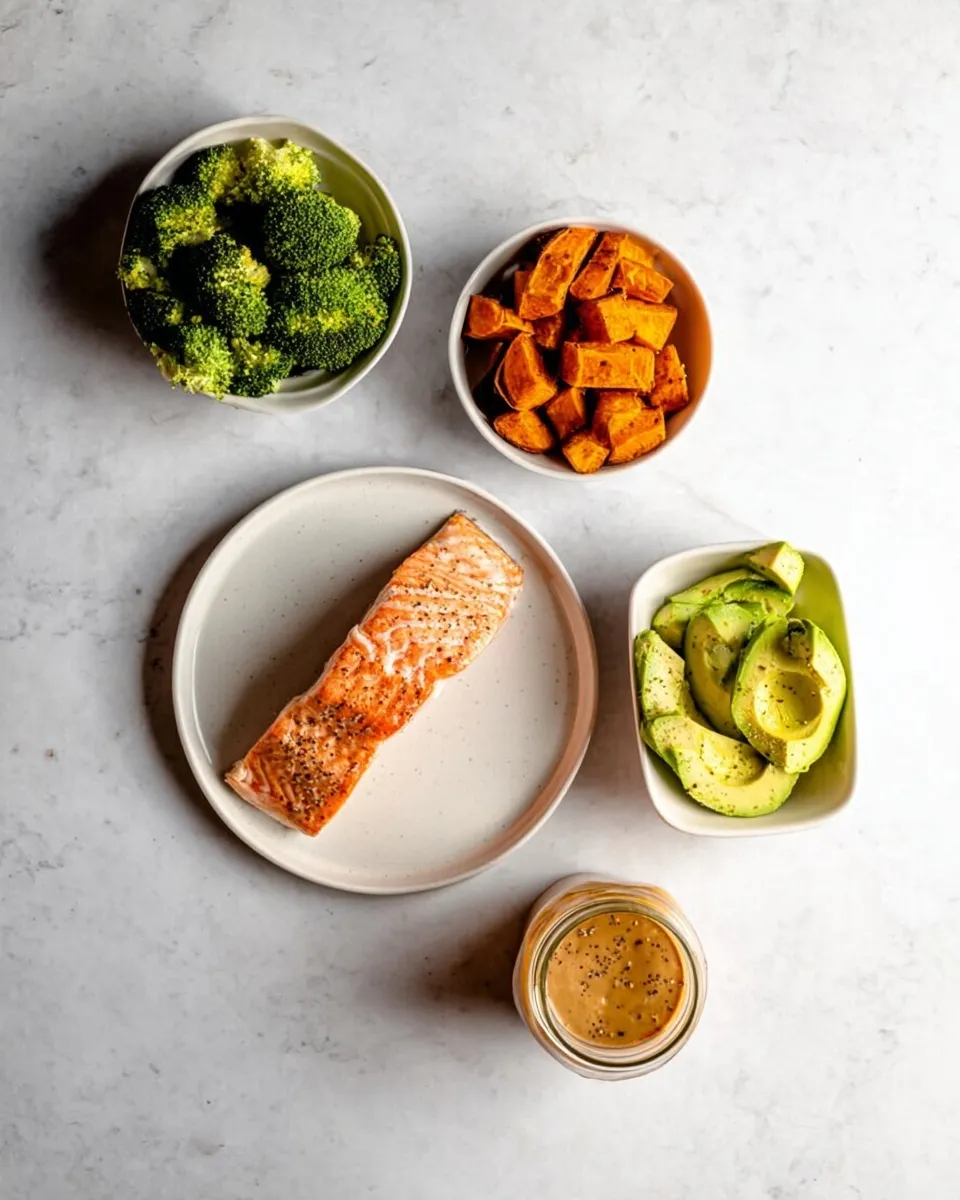 Flat lay photograph of healthy meal prep components in white ceramic bowls on a light speckled countertop. The ingredients include roasted broccoli florets, cubed roasted sweet potatoes, a perfectly seared salmon fillet, thinly sliced fresh avocado, and a small glass jar filled with a creamy, nutty sauce topped with seeds.