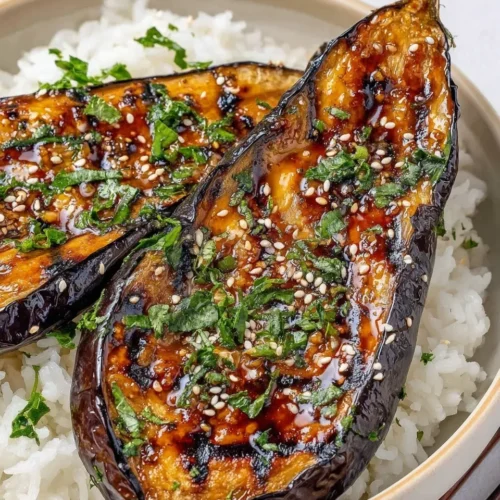 A close-up, high-angle shot of a Miso Glazed Eggplant rice bowl. Two halves of roasted eggplant are coated in a dark, glossy miso sauce with visible charred spots. They are served over fluffy white steamed rice and garnished generously with white sesame seeds and chopped green herbs. Wooden chopsticks are resting on the side of the ceramic bowl.