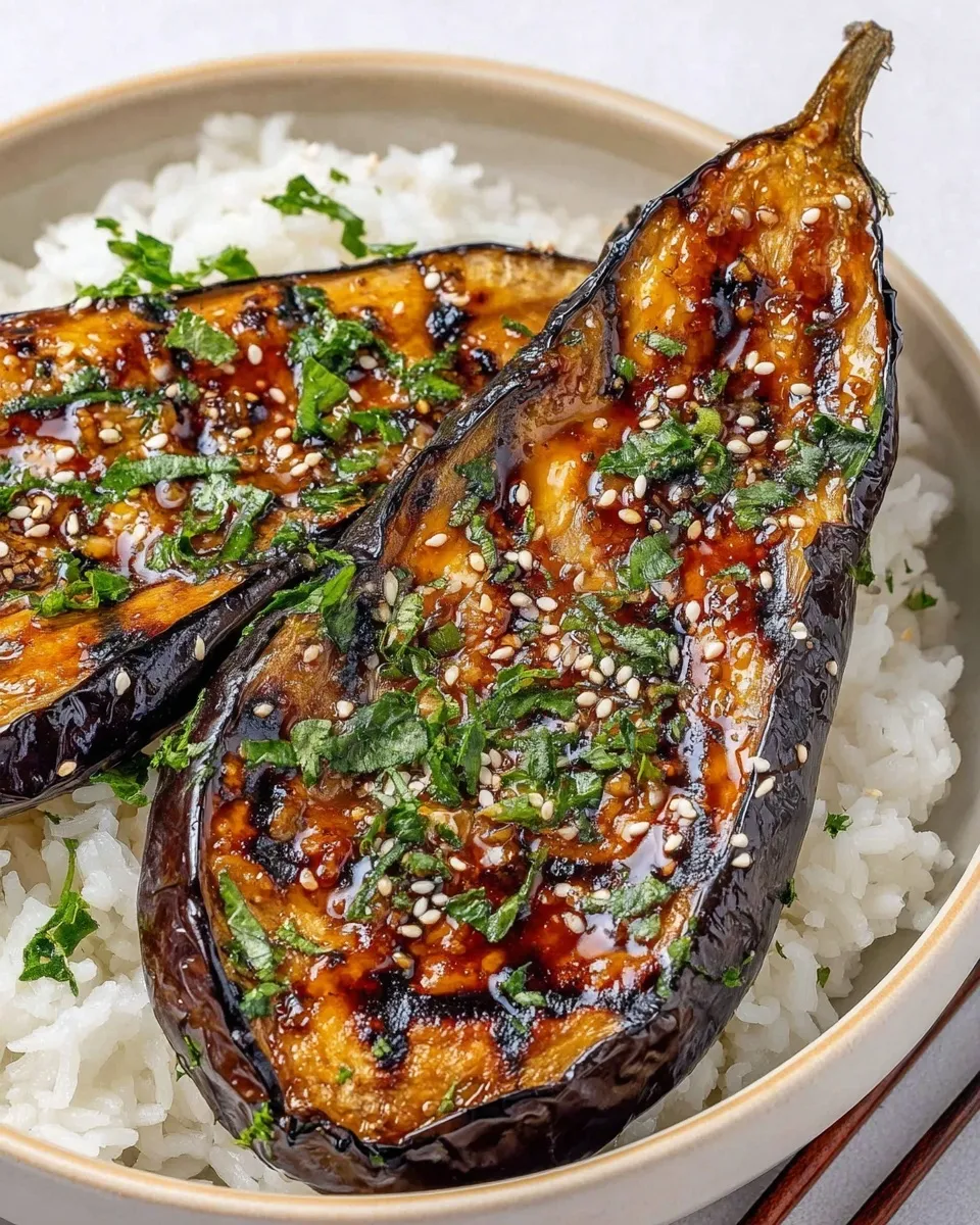 A close-up, high-angle shot of a Miso Glazed Eggplant rice bowl. Two halves of roasted eggplant are coated in a dark, glossy miso sauce with visible charred spots. They are served over fluffy white steamed rice and garnished generously with white sesame seeds and chopped green herbs. Wooden chopsticks are resting on the side of the ceramic bowl.