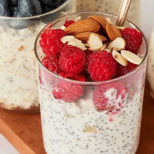 Close-up of a glass jar filled with creamy chia and protein overnight oats topped with fresh red raspberries and sliced almonds.