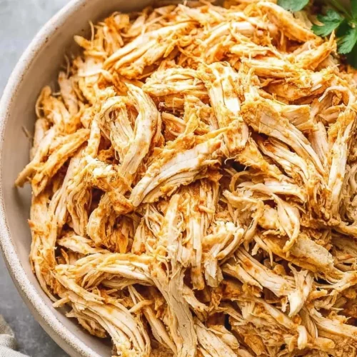 Close-up overhead view of tender, seasoned slow cooker shredded chicken in a beige ceramic bowl, garnished with a fresh parsley sprig.