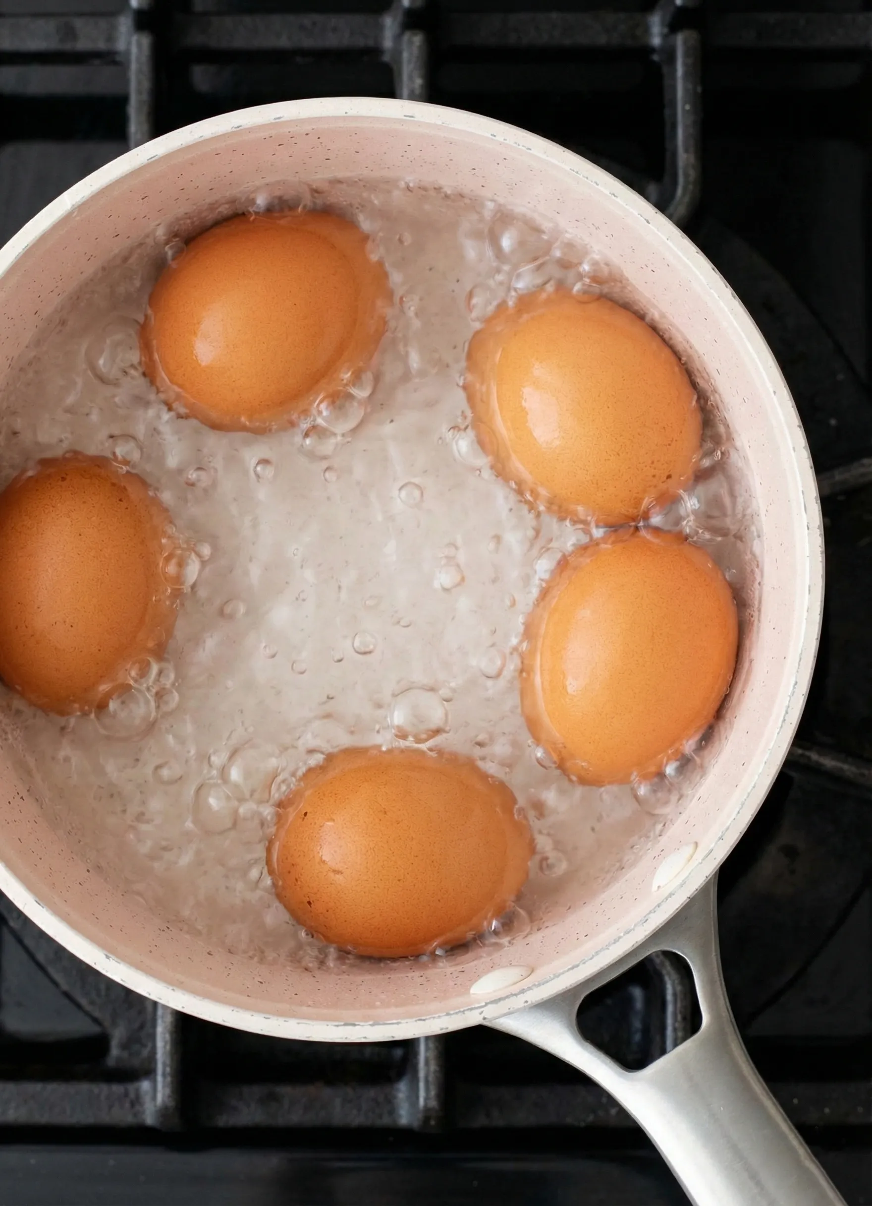 large brown eggs cooking Top-down view of five large brown eggs cooking in a light pink pot filled with vigorously boiling water on a black stovetop grate.