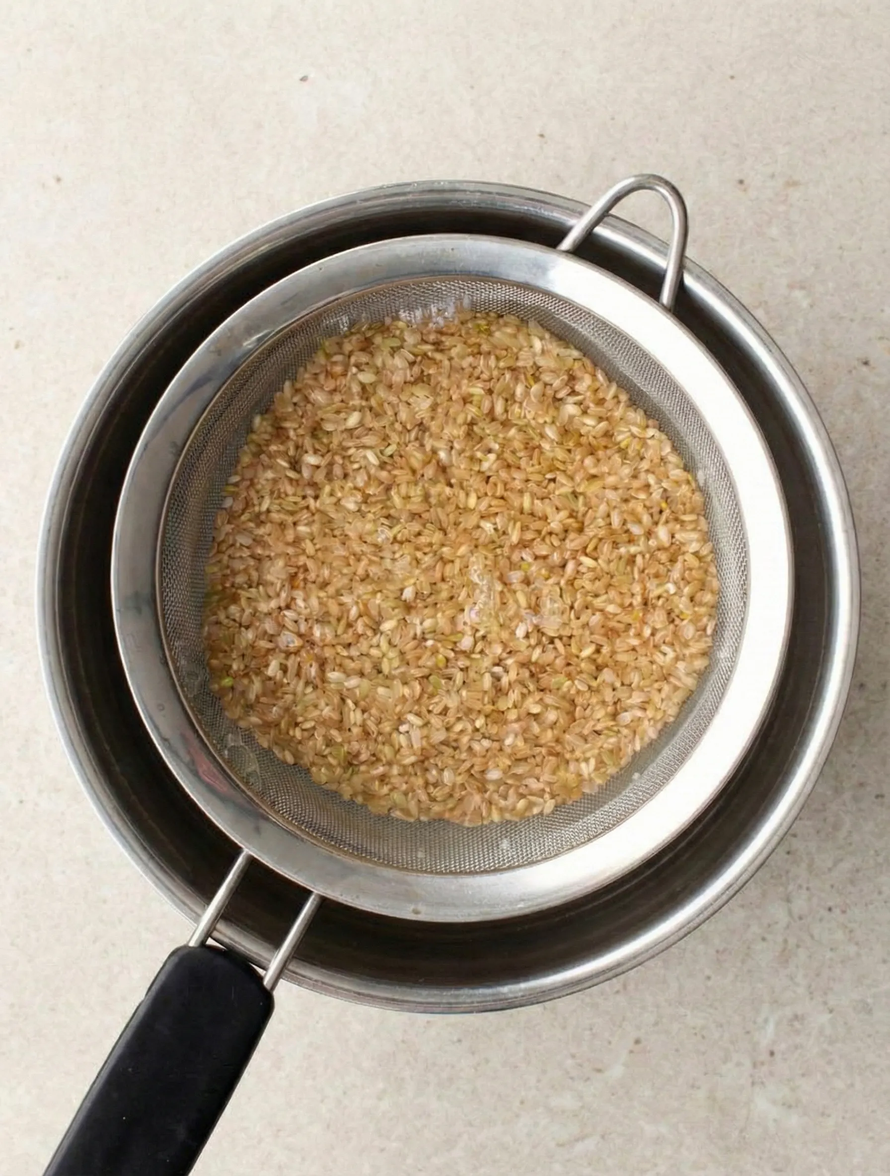 Overhead view of uncooked brown rice being rinsed in a stainless steel fine-mesh sieve set over a metal bowl to remove excess starch.