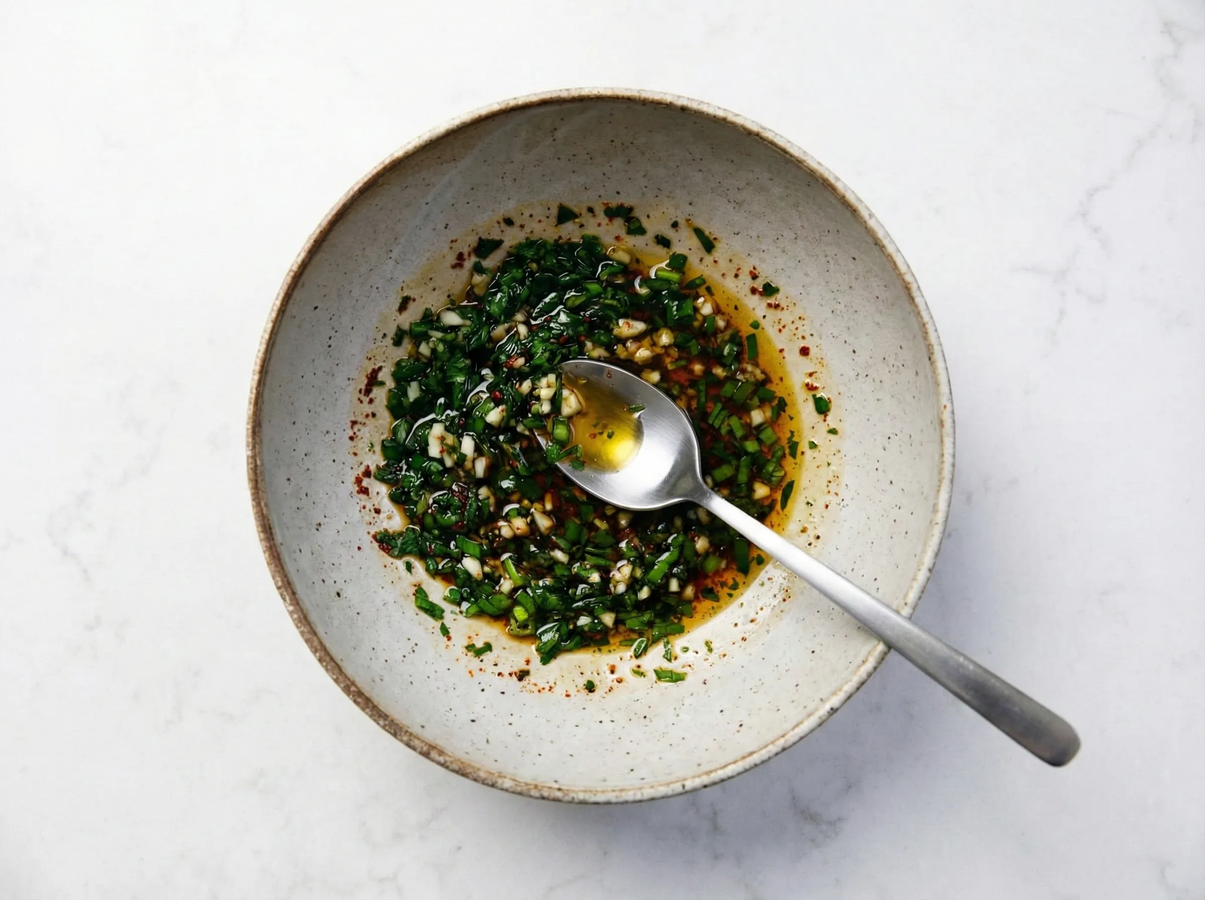 Overhead view of the aromatic chili oil sauce base in a speckled bowl, featuring chopped scallions, minced garlic, red chili flakes, and soy sauce mixed with oil, with a silver spoon resting inside.