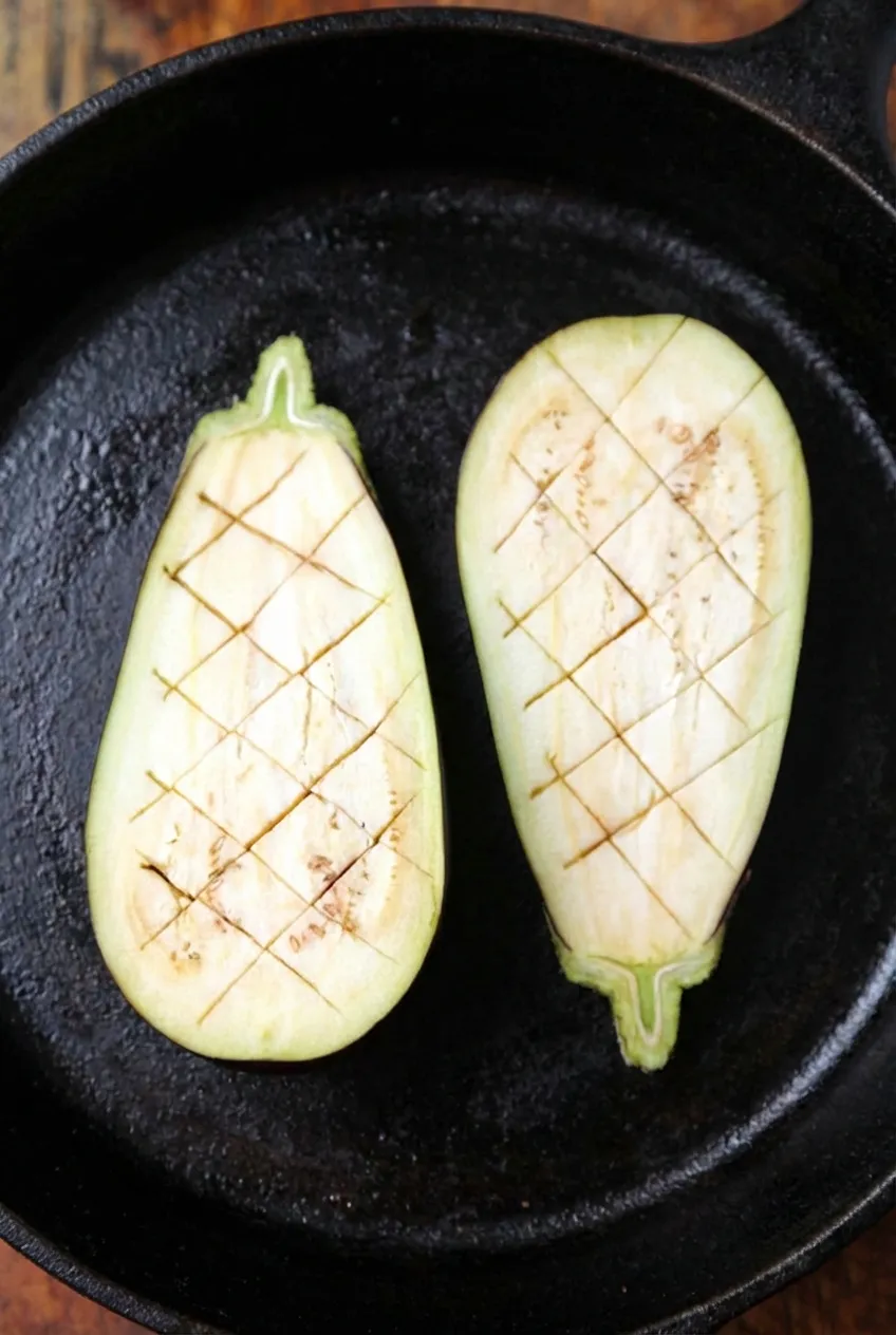 Top-down view of two raw eggplant halves placed in a black cast iron skillet. The white inner flesh is deeply scored with a diagonal crosshatch (diamond) pattern to prepare it for cooking.
