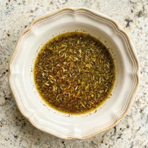 Overhead view of a beige ceramic bowl containing whisked Greek chicken marinade with olive oil, lemon juice, and visible dried herbs sitting on a speckled granite countertop.