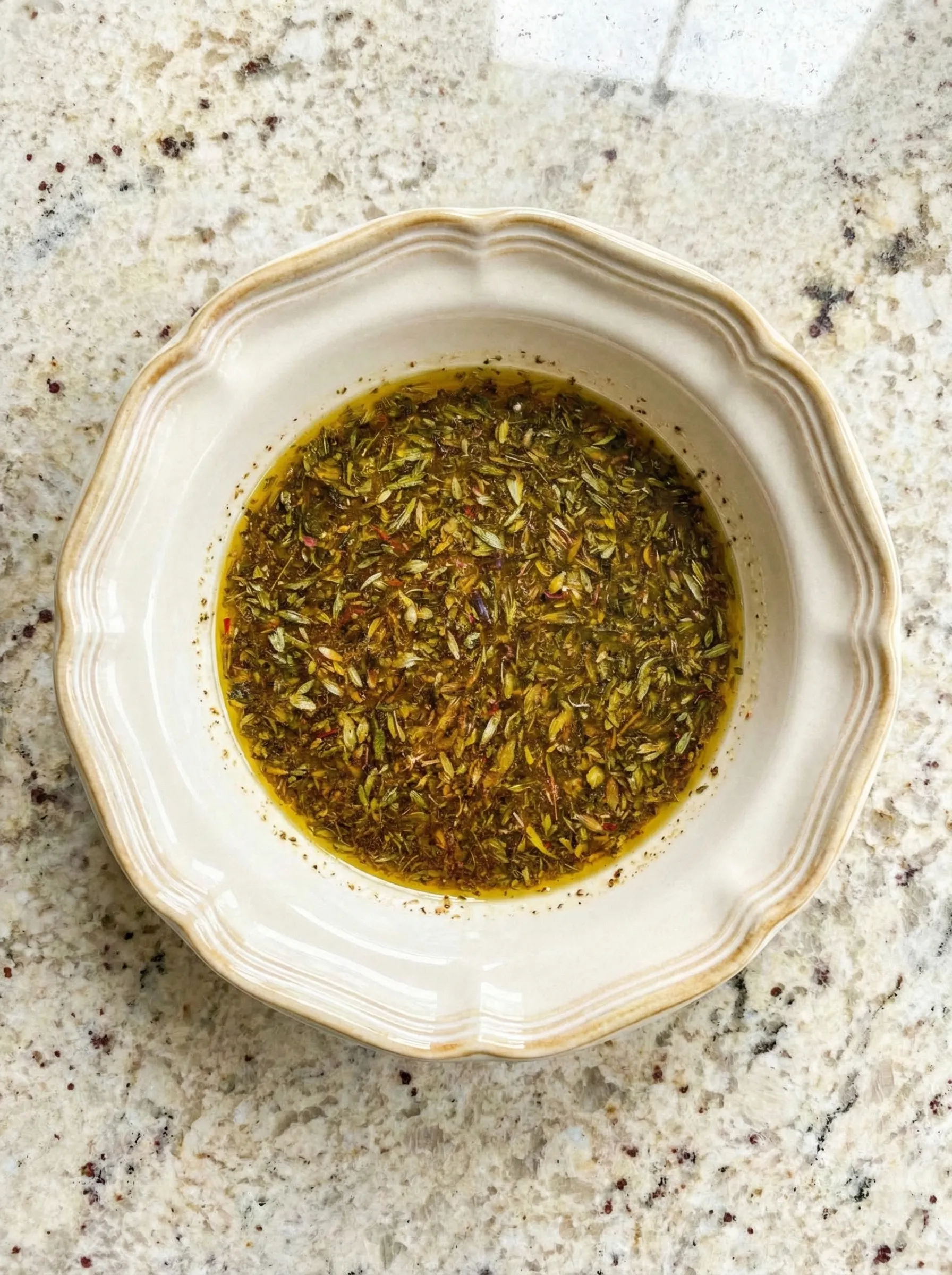 Overhead view of a beige ceramic bowl containing whisked Greek chicken marinade with olive oil, lemon juice, and visible dried herbs sitting on a speckled granite countertop.