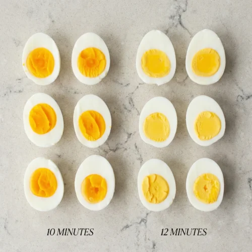 Top-down comparison of halved hard boiled eggs arranged in a grid, showing the difference between the creamy, vibrant orange yolks of 10-minute eggs versus the firm, pale yellow yolks of 12-minute eggs.