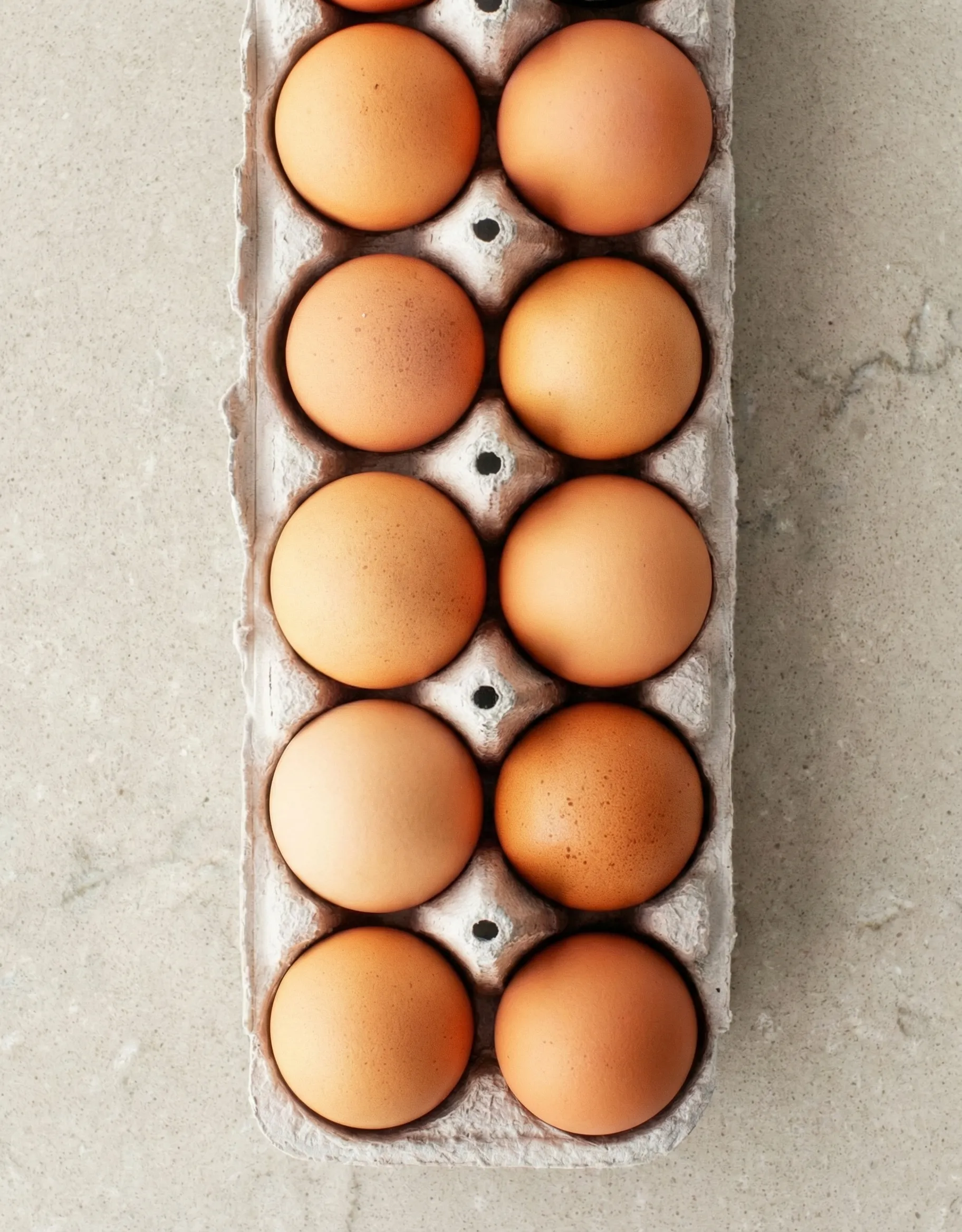 ingredients Top-down view of an open cardboard carton filled with large brown eggs on a neutral stone countertop, the main ingredient for making perfect hard boiled eggs.