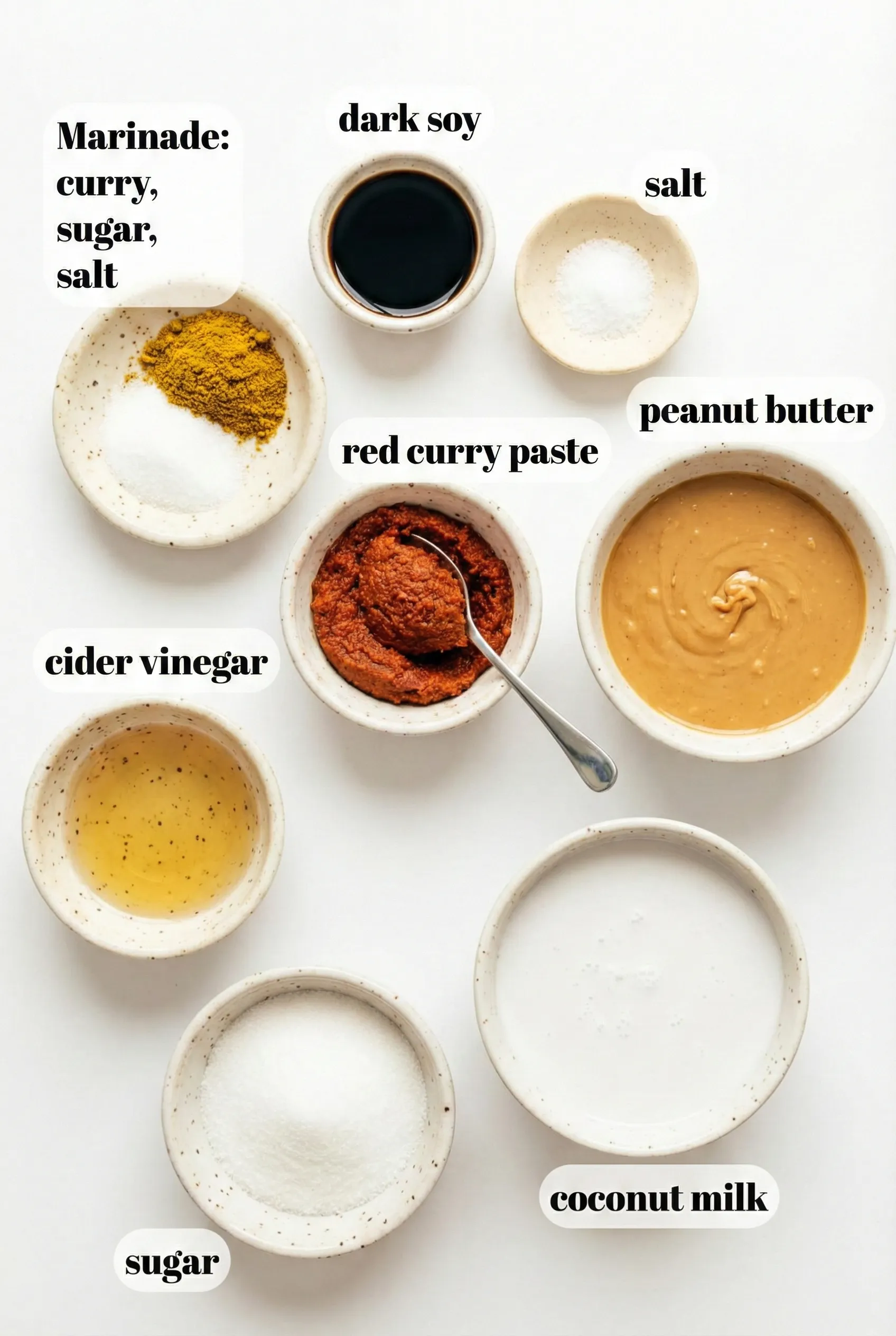 A flat lay arrangement of ingredients for Thai peanut chicken in small ceramic bowls on a white surface. Text labels identify marinade spices (curry, sugar, salt), dark soy sauce, red curry paste, peanut butter, cider vinegar, sugar, and coconut milk.